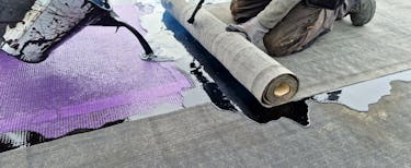 A worker installing roofing material kneels on a flat surface while unrolling a large roll of grey roofing felt. Liquid black adhesive is being poured onto the surface, with a grid-like purple layer partially visible underneath. The worker is wearing protective gloves and trousers.